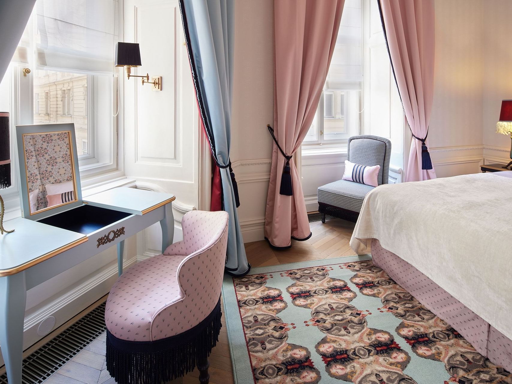 Elegant hotel bedroom with large bed, patterned rug, vanity, chair, and windows with pink curtains.