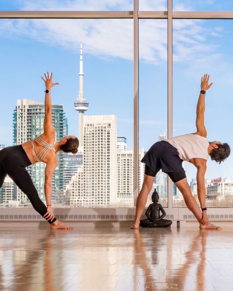 Two people practicing yoga in a side bend pose with Toronto skyline and CN Tower visible through floor-to-ceiling windows