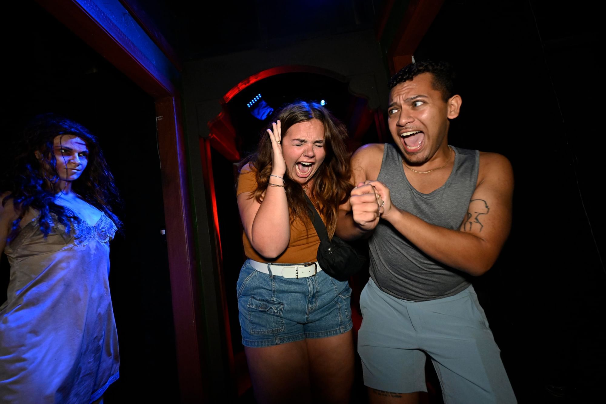 A man and a woman hold hands and scream in terror as they pass a creepy woman in a nightgown in a dark hallway at Universal’s Halloween Horror Nights, an amazing Halloween event in Orlando.