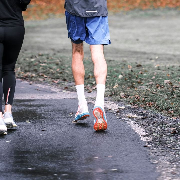 Zwei Personen joggen auf einem nassen Pfad, umgeben von Herbstlaub und Gras.