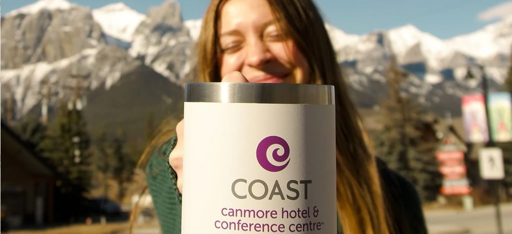 A person holds out a Coast Canmore Hotel & Conference Centre mug on a sunny day in front of the mountains.