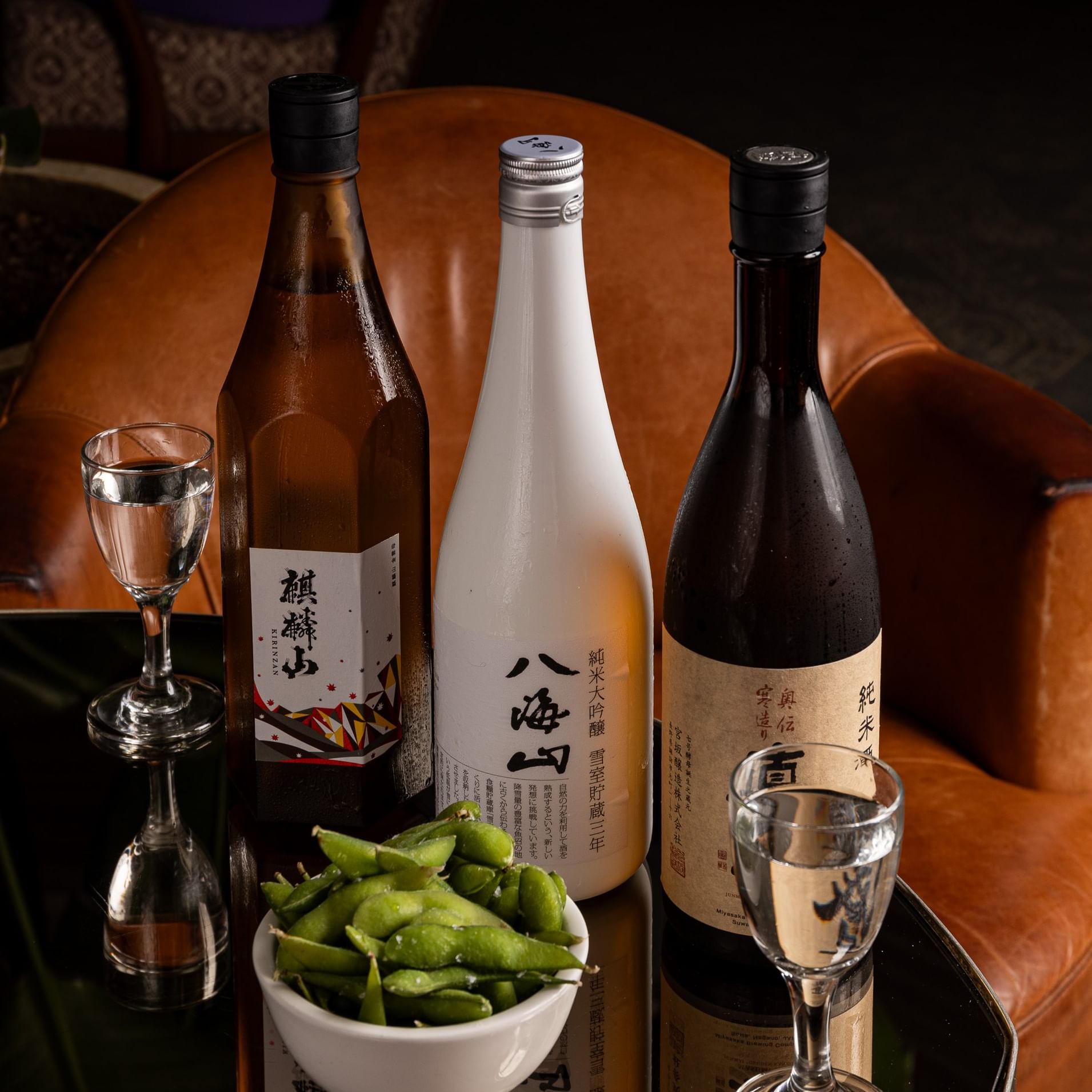 Three sake bottles with glasses and edamame displayed on a leather chair for National Sake Day at Blue Ribbon.