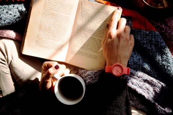 An aerial view of someone holding a cup of coffee and reading an open book in their lap.