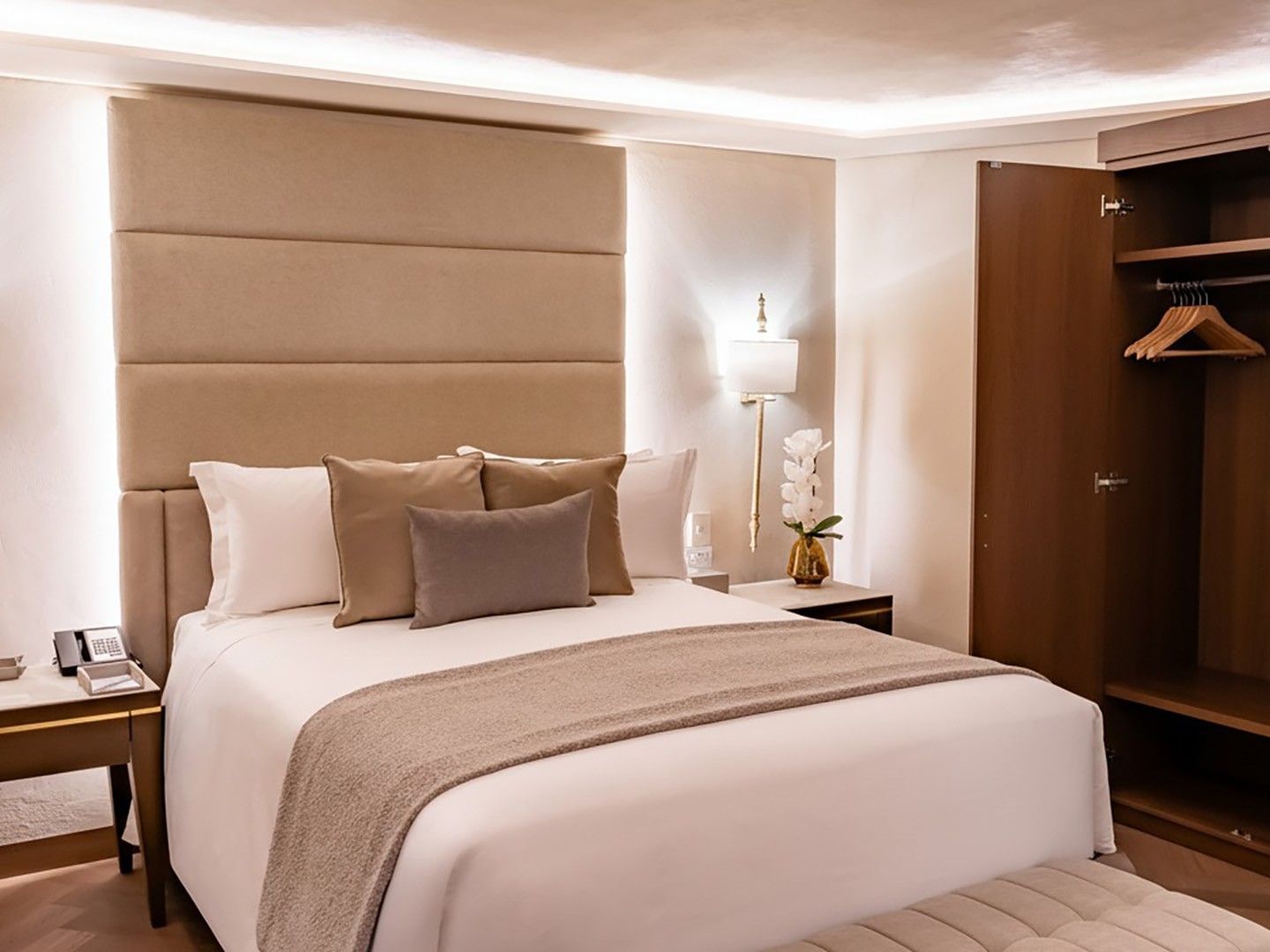 Modern hotel bed with a tall tan headboard, white linens, and an open wooden wardrobe in Queen Novicia at Quinta Real Puebla