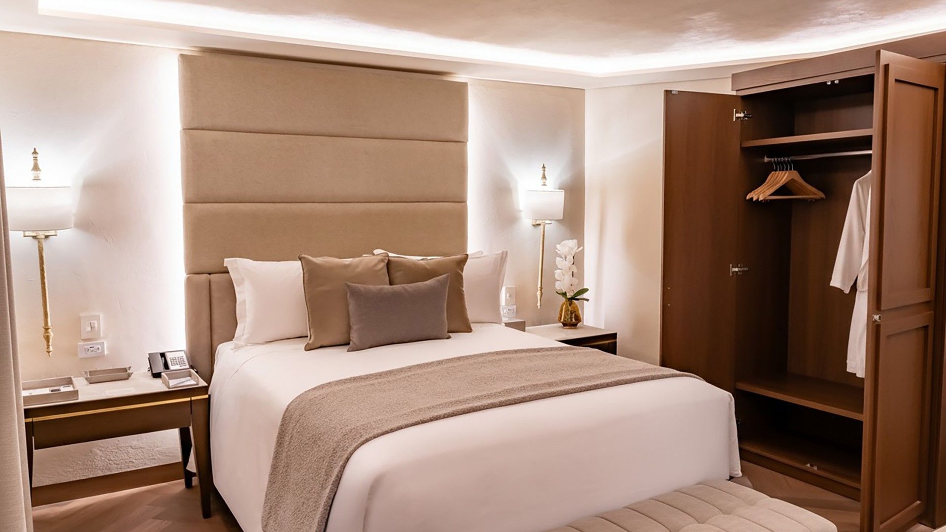 Modern hotel bed with a tall tan headboard, white linens, and an open wooden wardrobe in Queen Novicia at Quinta Real Puebla