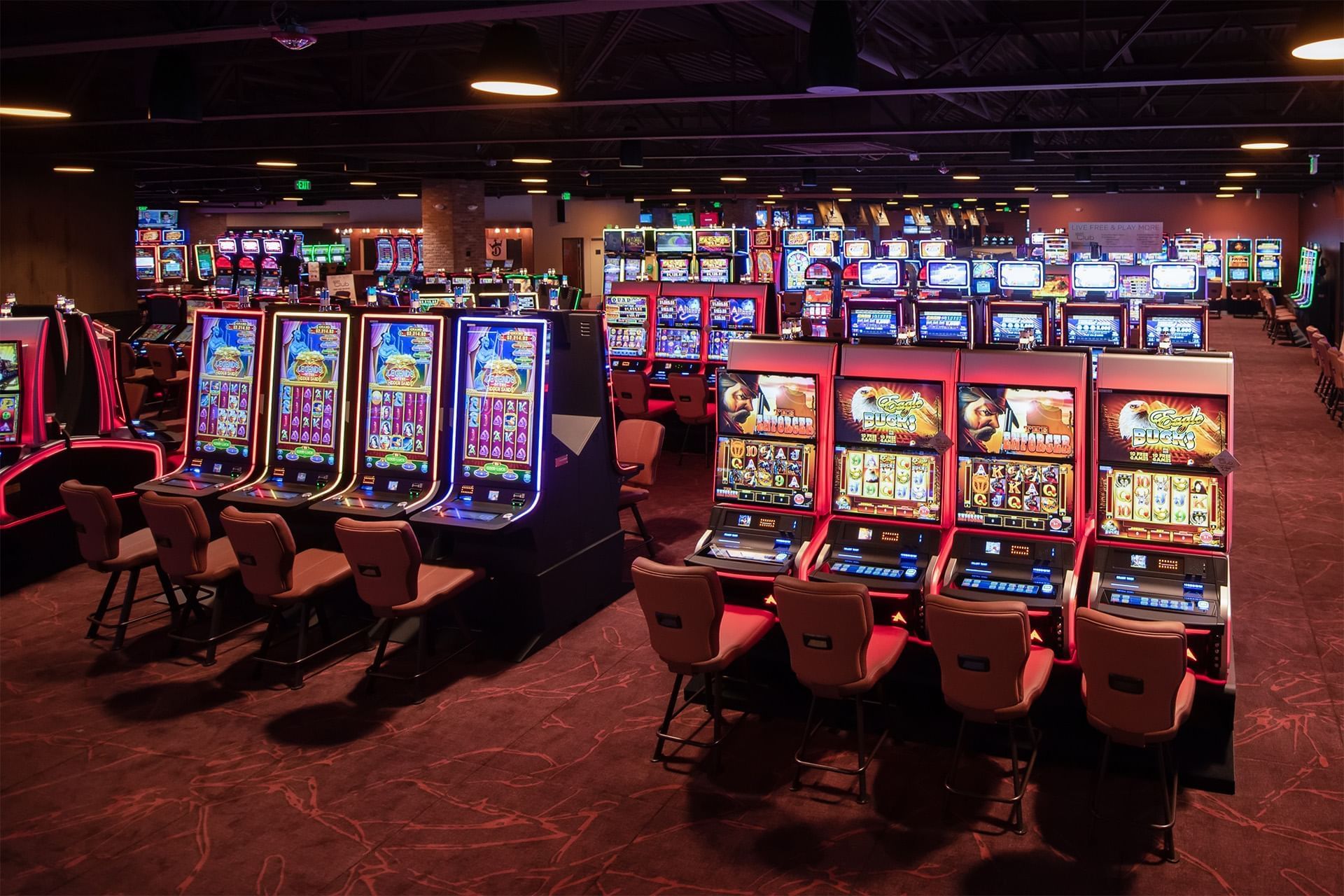 Rows of casino gaming machines with padded chairs arranged across the carpeted floor under ceiling lights at The Brook