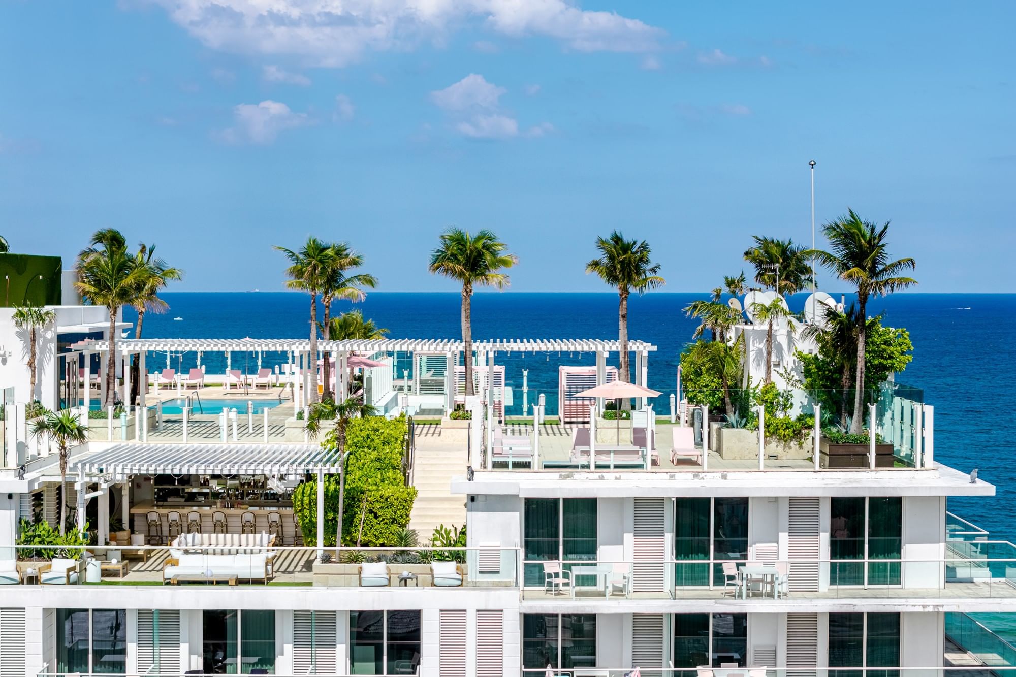 A white multi-story building with a pool and palm trees on the roof, overlooking the ocean.