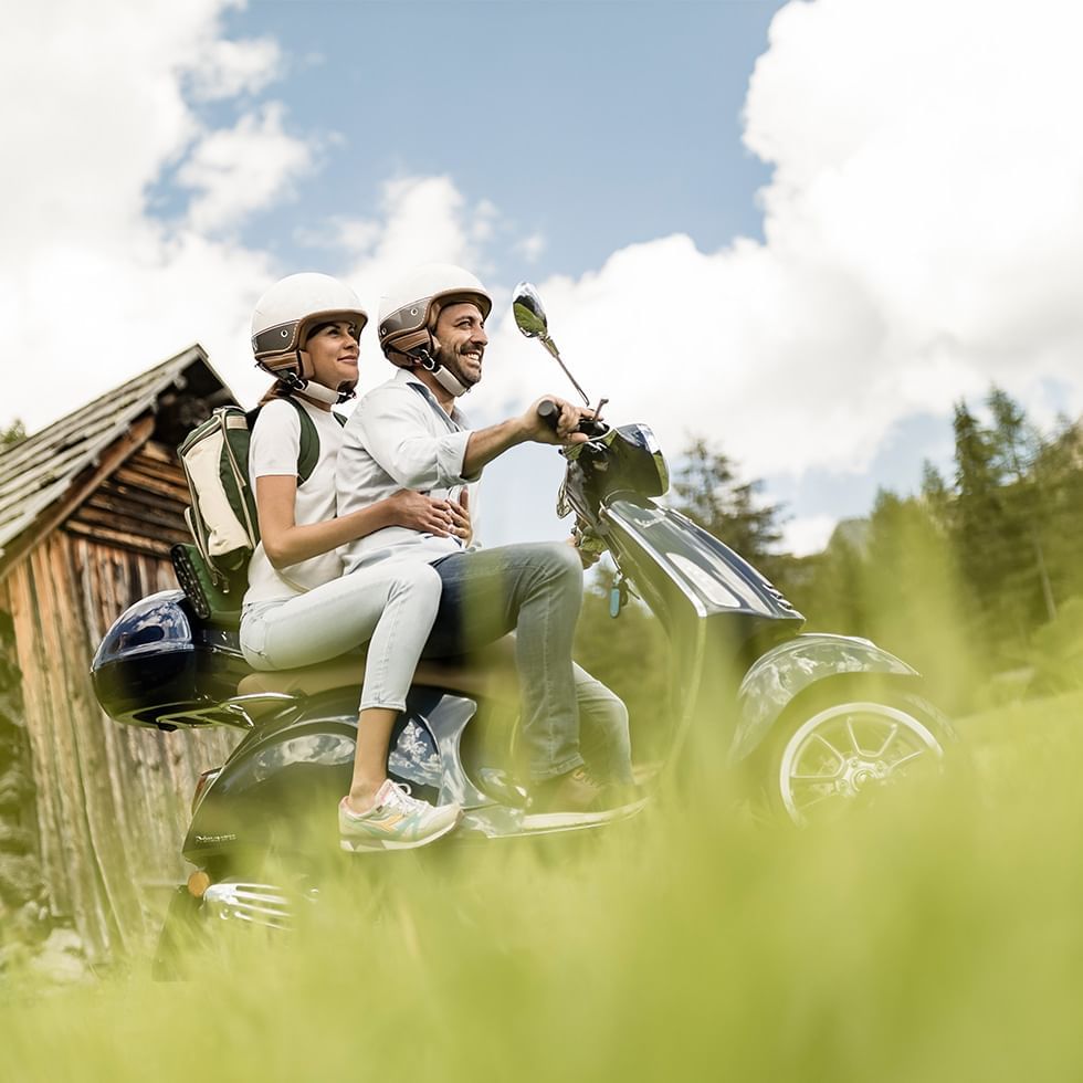 Couple on a scooter bike by a wooden cabin Falkensteiner Hotels