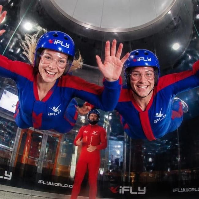 Two smiling participants in blue iFLY suits and helmets floating in an indoor skydiving wind tunnel with an instructor below.