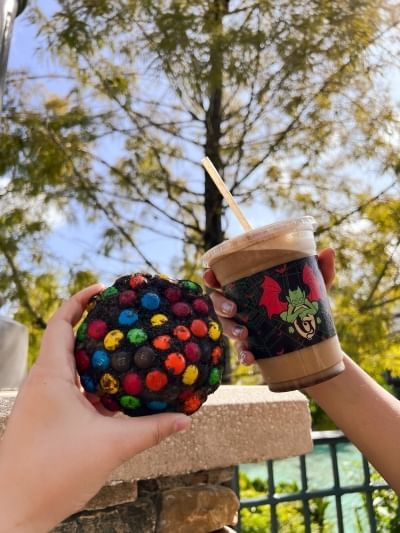 Two hands hold a large chocolate cookie with colorful candies and an iced coffee from Gideon's Bakehouse, a bakery in Orlando.