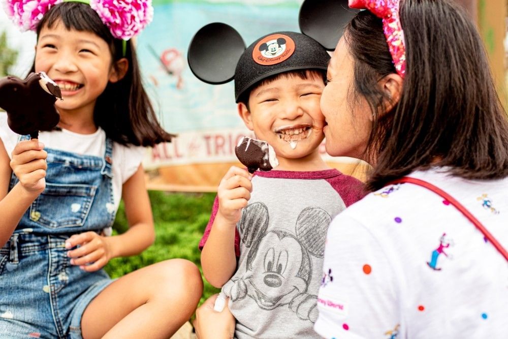 A boy and girl in Mickey and Minnie attire eat Mickey ice cream bars, while their mother kisses the boy on the cheek.