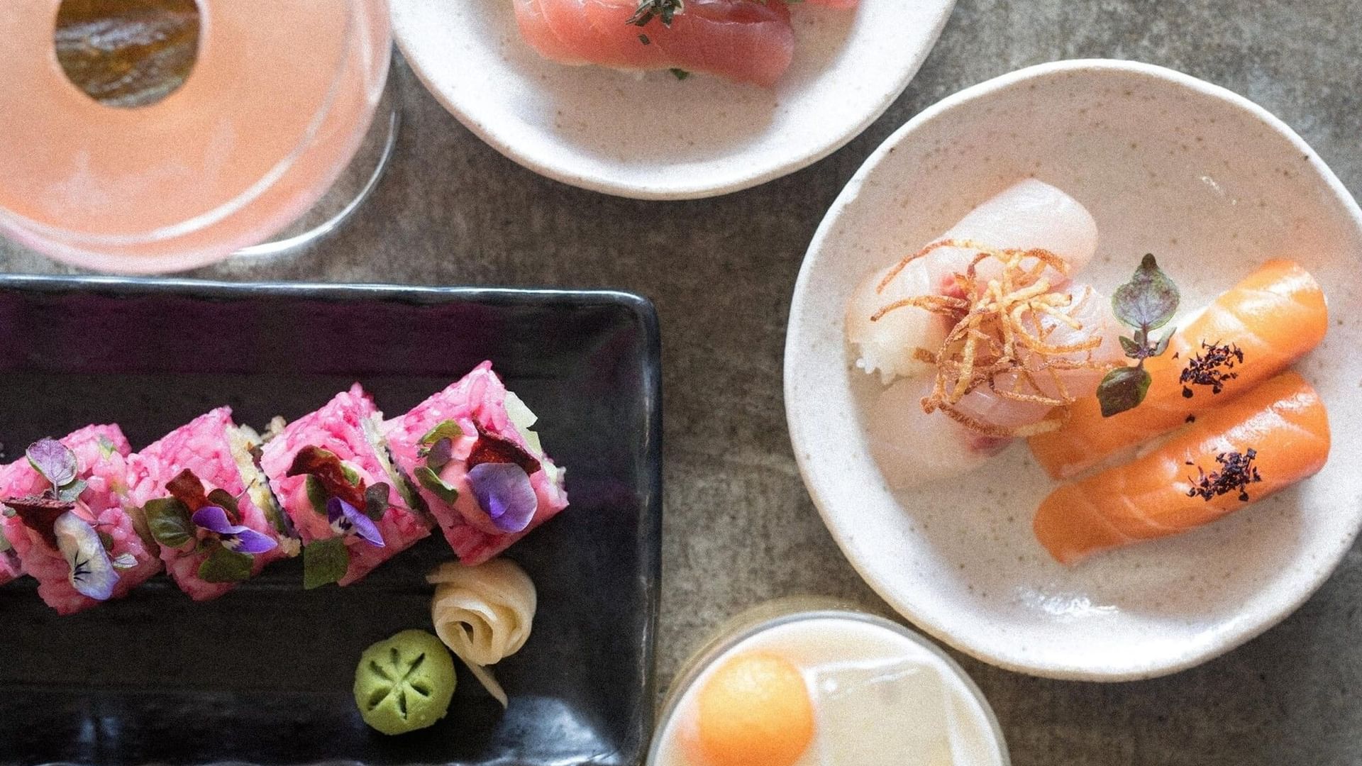 Sunset Sabi with plates of fresh sushi arranged by cocktail drinks on a stone table under bright lights at Elysium Manly