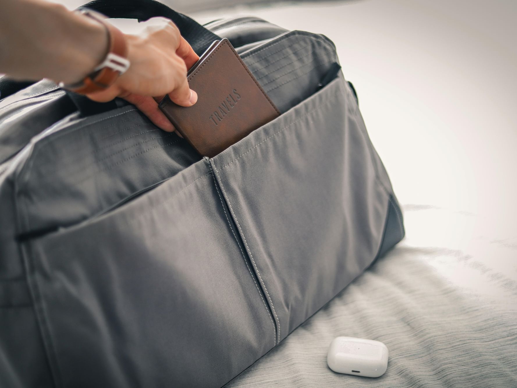 A close-up shot of a traveller's hand placing a brown leather travel wallet into a gray duffel bag on a bed, representing the Corporate Saver package at Hotel Grand Chancellor Townsville.