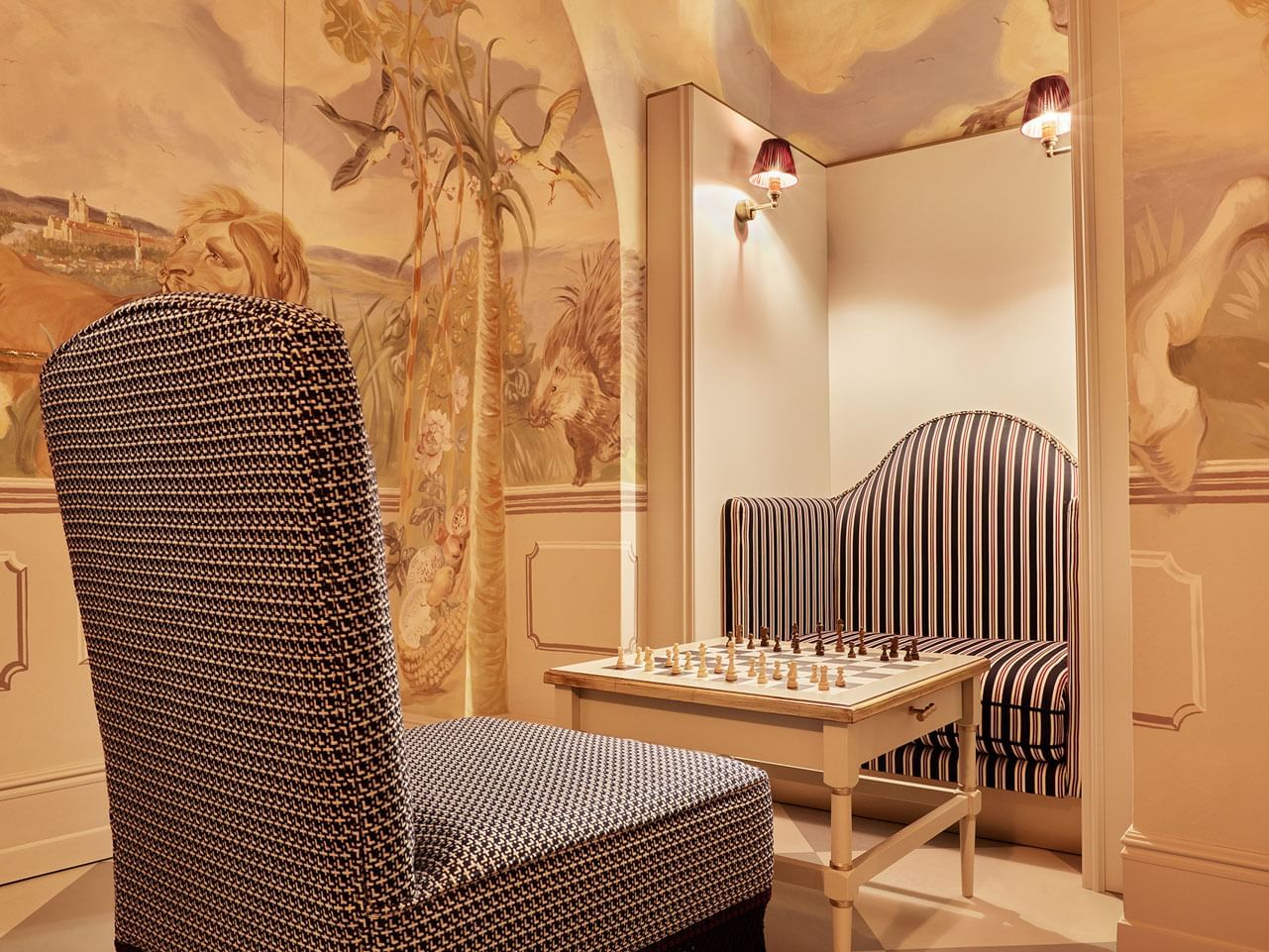 Hotel lobby with patterned chair, striped bench, chess table, and mural wall featuring pastoral scenes and wildlife.