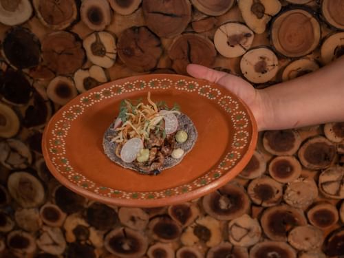 Hand holding a blue corn tortilla with grilled meat and radish slices on a plate in Tahona at Hacienda del Mar Los Cabos.