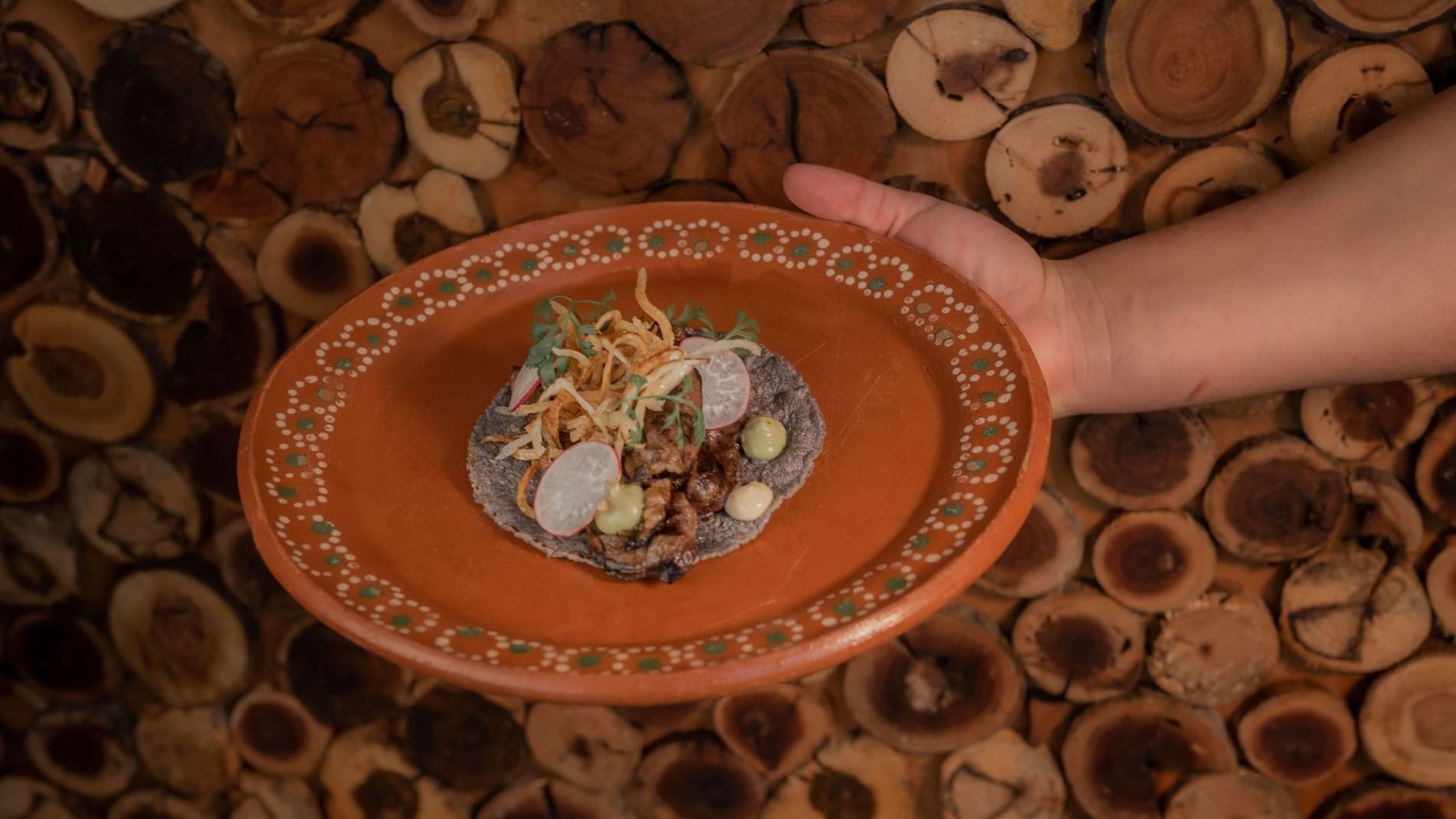 Hand holding a blue corn tortilla with grilled meat and radish slices on a plate in Tahona at Hacienda del Mar Los Cabos.