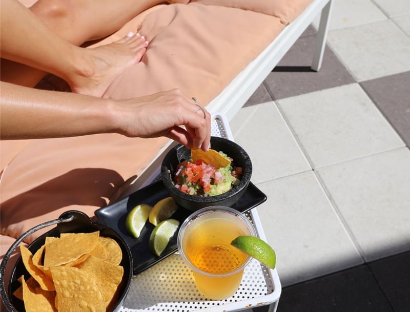 A person's hands holding a tortilla chip with guacamole and a drink on a tray