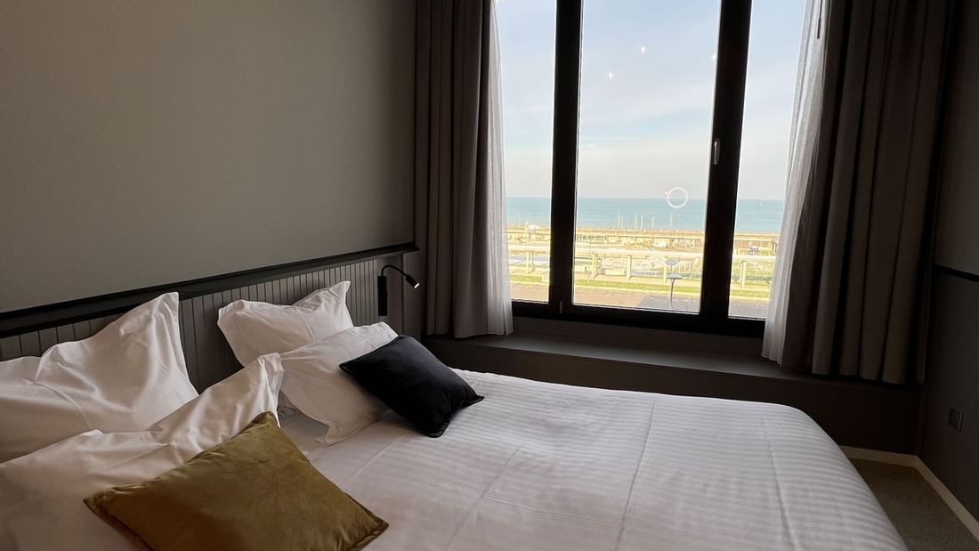 Chambre avec lit blanc, oreillers, et vue sur la mer par une grande fenêtre.