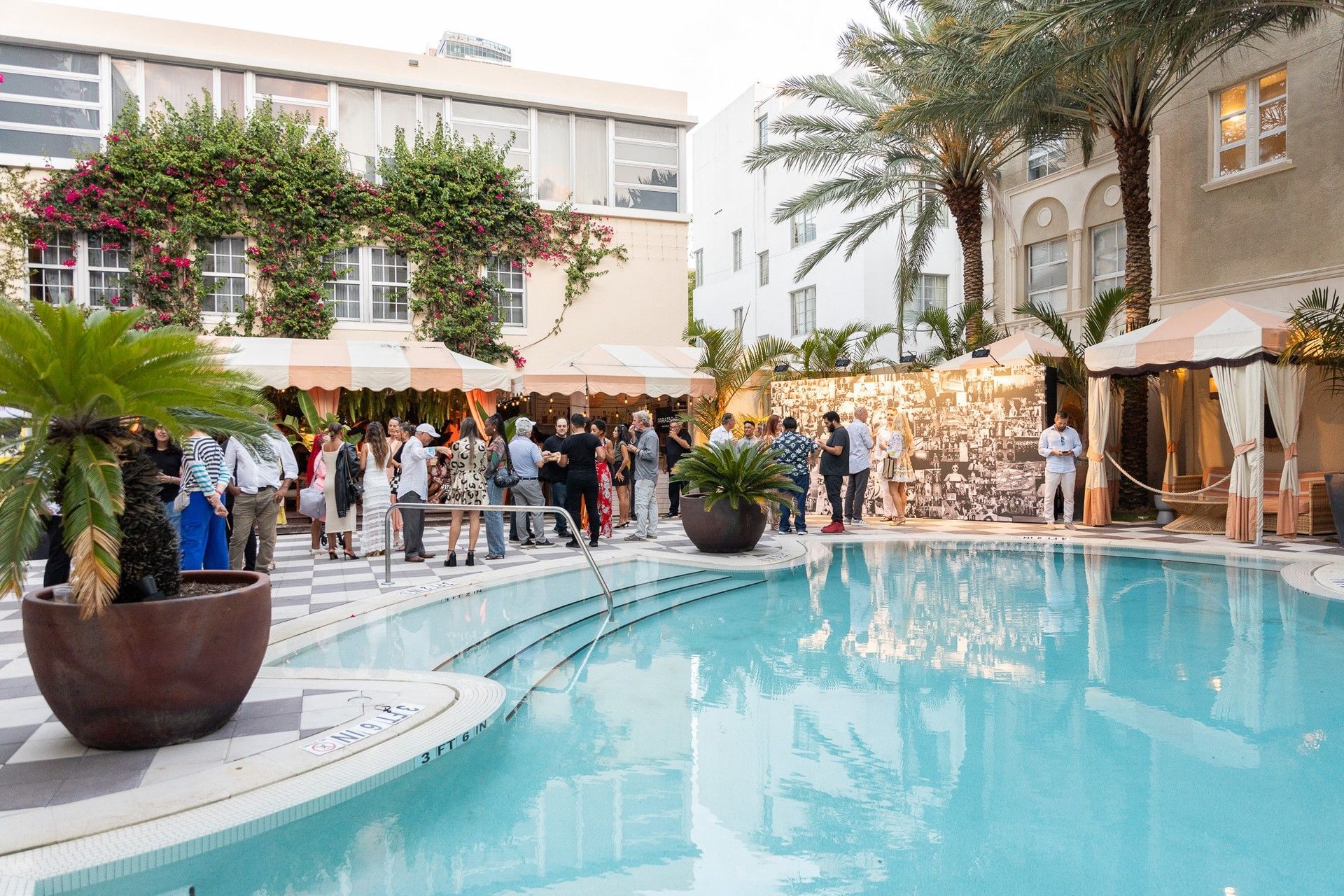 A group of people gathered around a pool with palm trees and umbrellas in the background.
