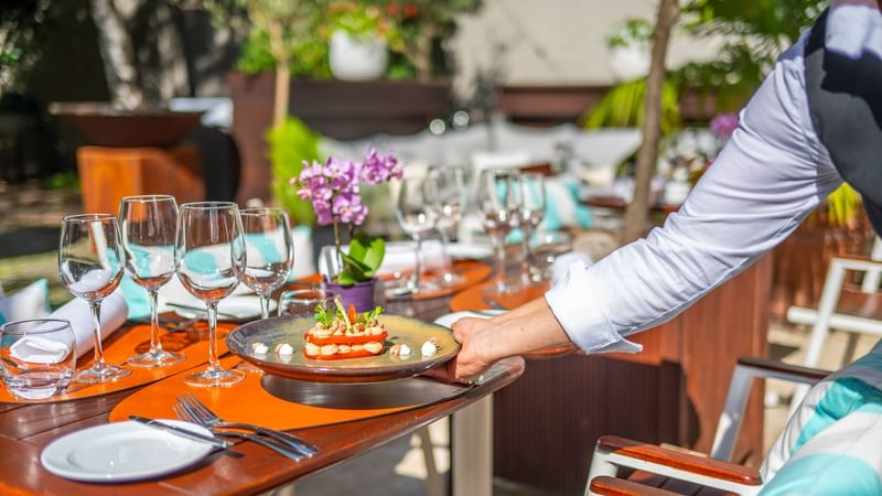 Waiter serves a gourmet dish at an outdoor table with wine glasses and tropical decor in Hotel Barsey by Warwick