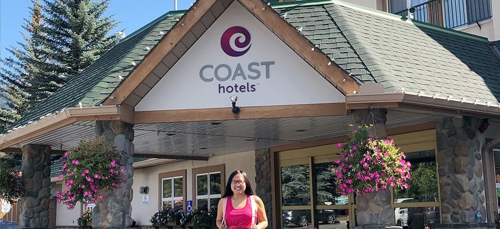 Woman standing outside the entrance of Coast Canmore Hotel with flower baskets hanging from stone pillars.