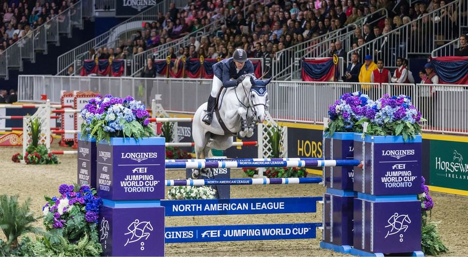 White horse with rider jumping over blue obstacle hurdle during show jumping competition event near Hotel X Toronto