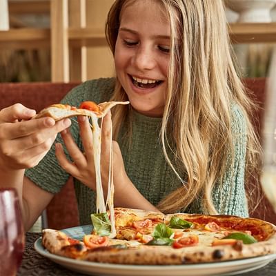 Mädchen mit blonden Haaren isst eine Pizza mit Käse und Tomaten auf einem Teller.