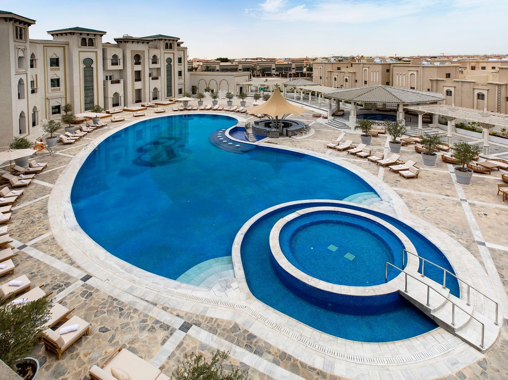 Large, intricately designed resort pool with lounge chairs and umbrellas, surrounded by elegant architecture.