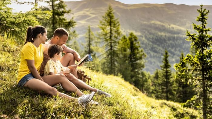 Familie liest gemeinsam ein Buch in einer bergigen Landschaft mit Bäumen und Gras.