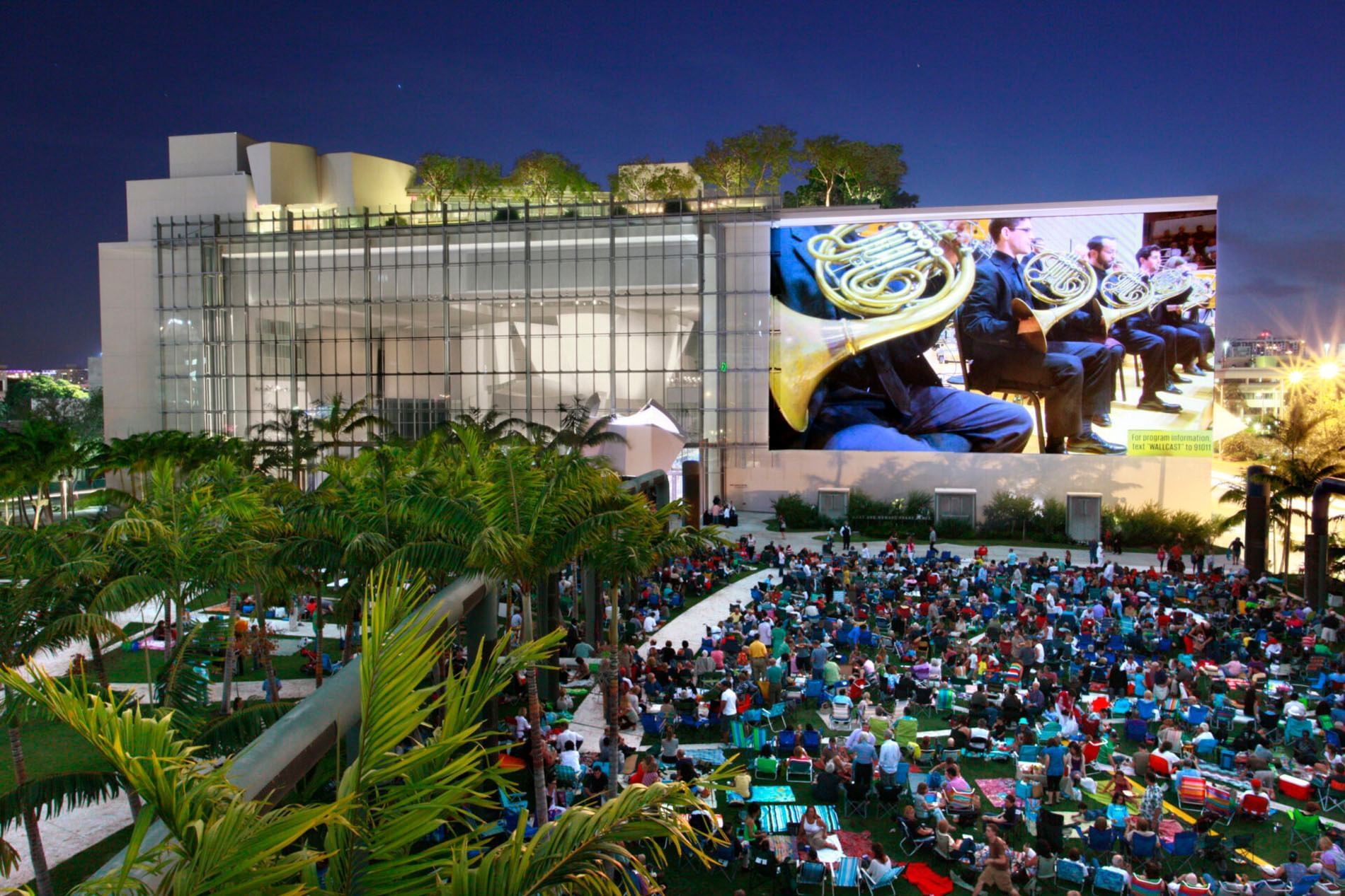 A large building with a screen on the side showing an image of musicians, with a crowd of people sitting in front of it.