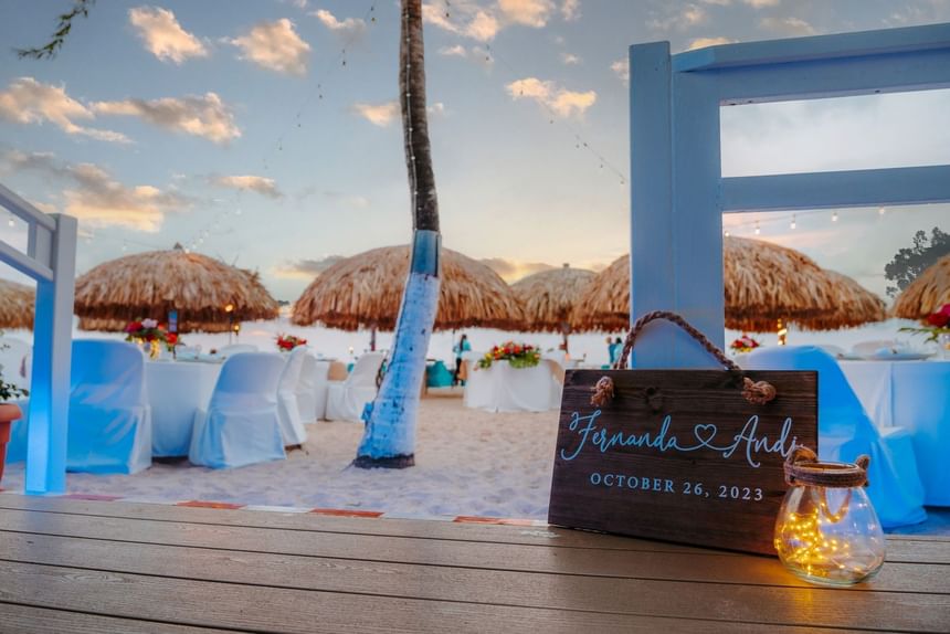 A beach wedding setup with blue chairs, white tables, wooden sign for Fernanda and Andi, and string lights.