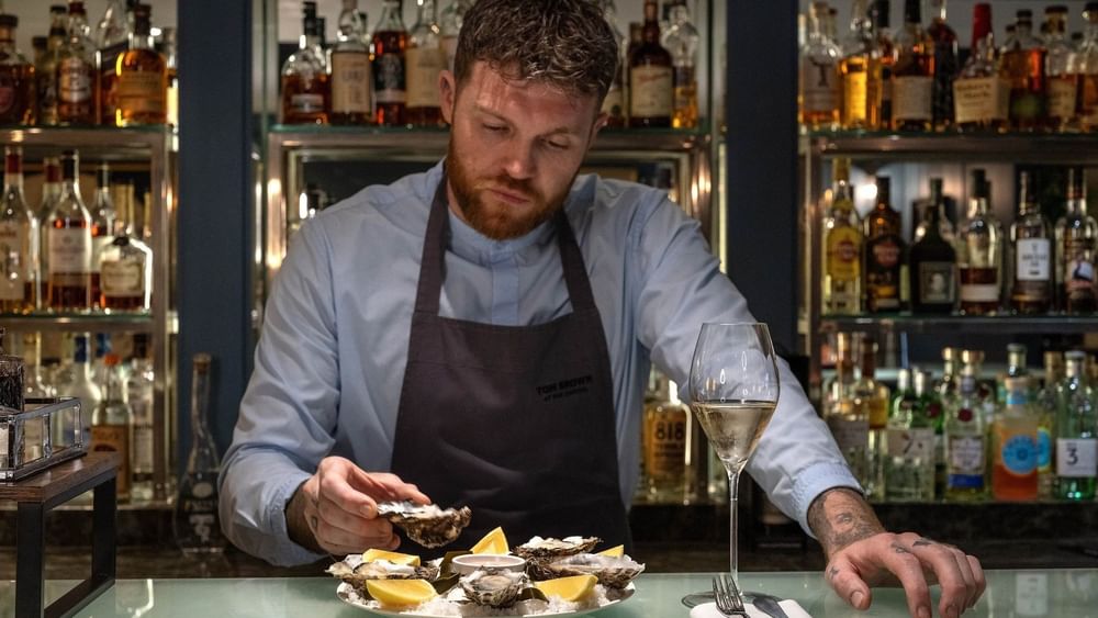 Bartender in a dark apron by shelves of liquor, preparing fresh oysters at The Capital Hotel, Apartments and Townhouse