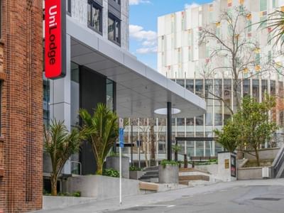 Modern urban entrance with red UniLodge signage, landscaped courtyard, and contemporary architecture at UniLodge Auckland Central.