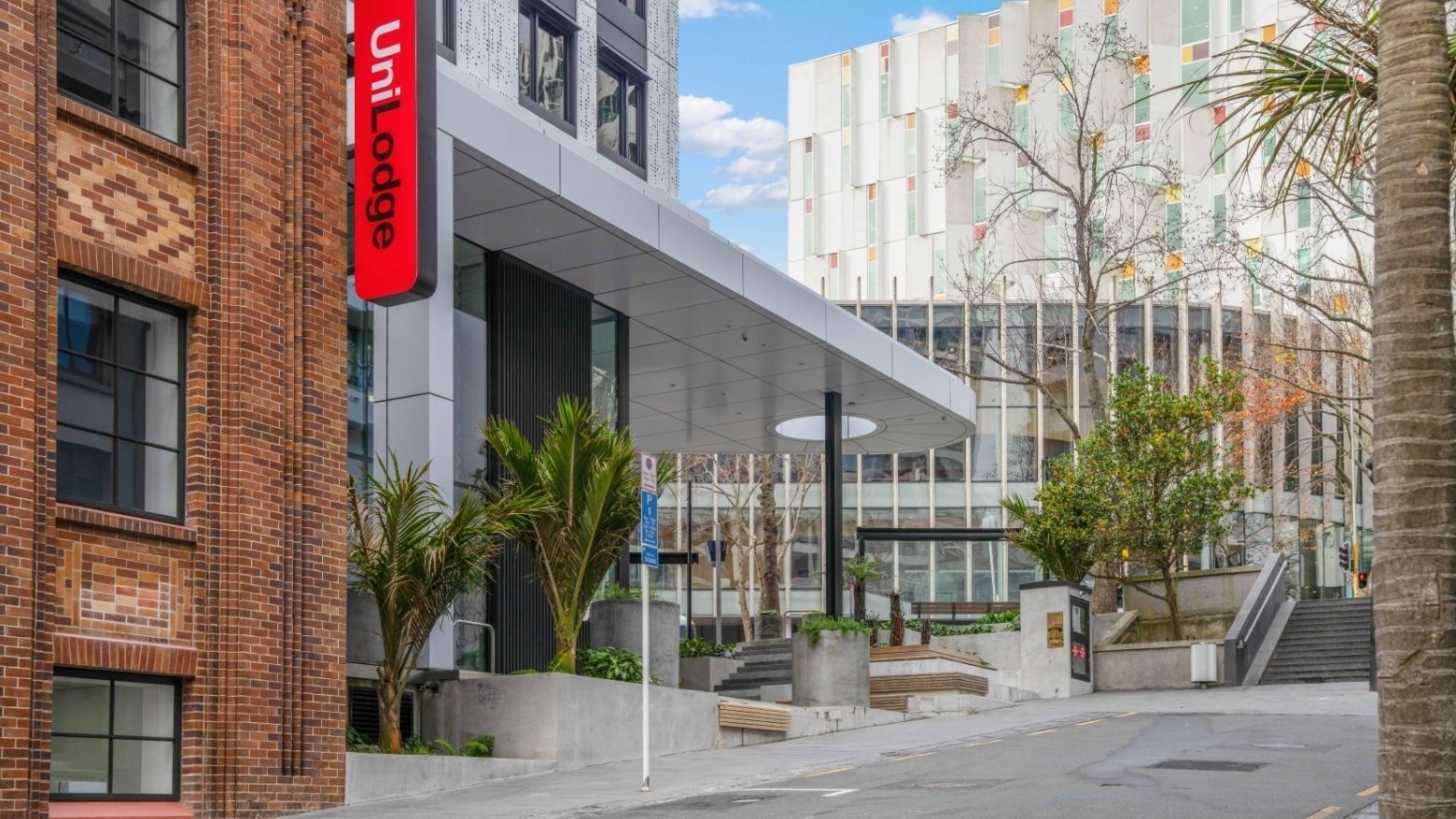 Modern urban entrance with red UniLodge signage, landscaped courtyard, and contemporary architecture at UniLodge Auckland Central.