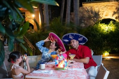 A family of three wearing sombreros sit at a table outside at night, with one child taking a photo.