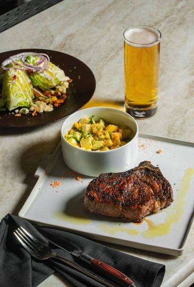 Seared steak on a white plate, accompanied by a salad, a bowl of diced vegetables, and a glass of beer at Cypress & Oak