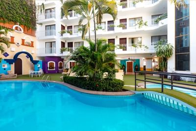 Piscina curva rodeada de palmeras y jardines con edificio blanco de balcones al fondo en Los Arcos Suites