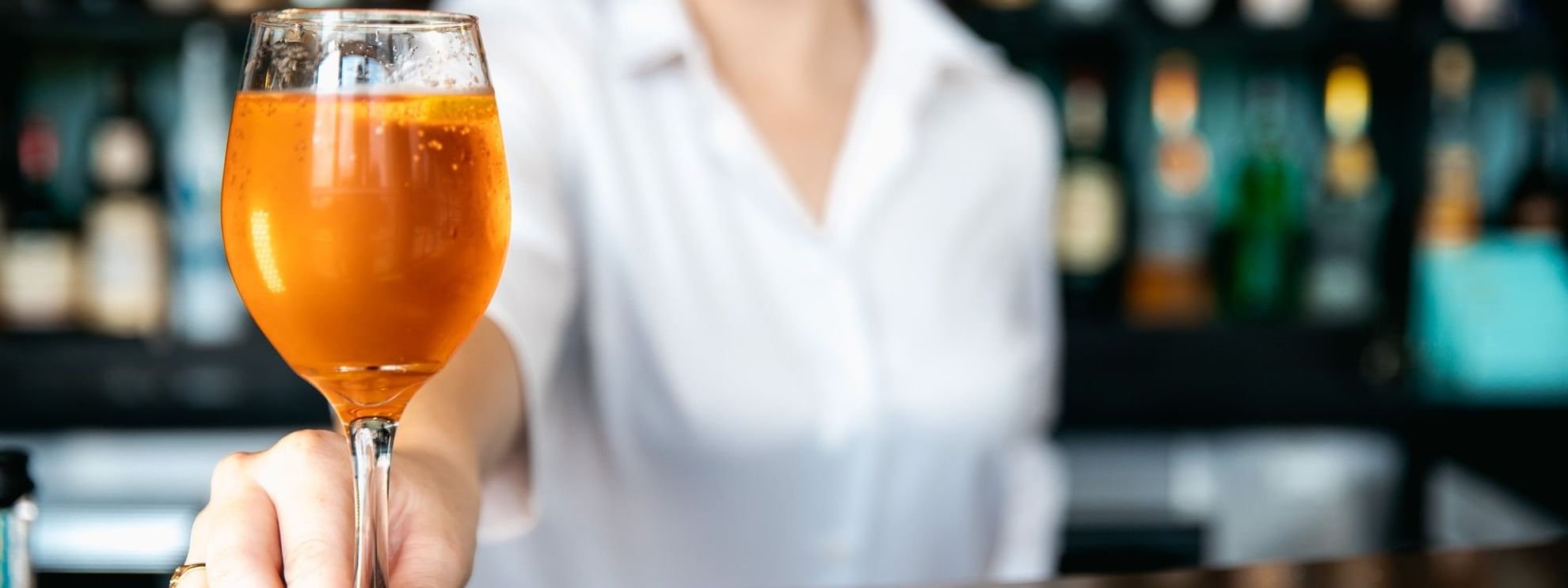 Woman holds a glass of orange beverage at a bar with blurred bottles in background.
