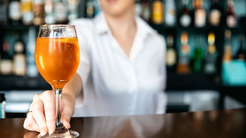 Woman holds a glass of orange beverage at a bar with blurred bottles in background.