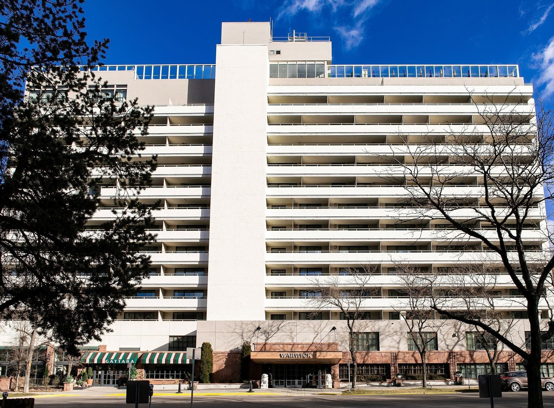 The Warwick Hotel Denver high-rise exterior, near the Gattara sign, framed by pine and leafless trees