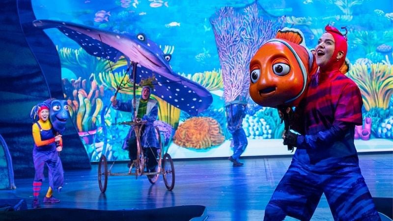 Performers in colorful costumes carry elaborate puppets of characters from Finding Nemo on a stage with an underwater backdrop.
