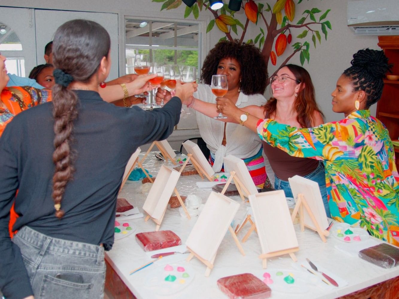 Group of women toasting with drinks at a creative workshop table with art supplies and cacao pods at Cocoa By The Bay.