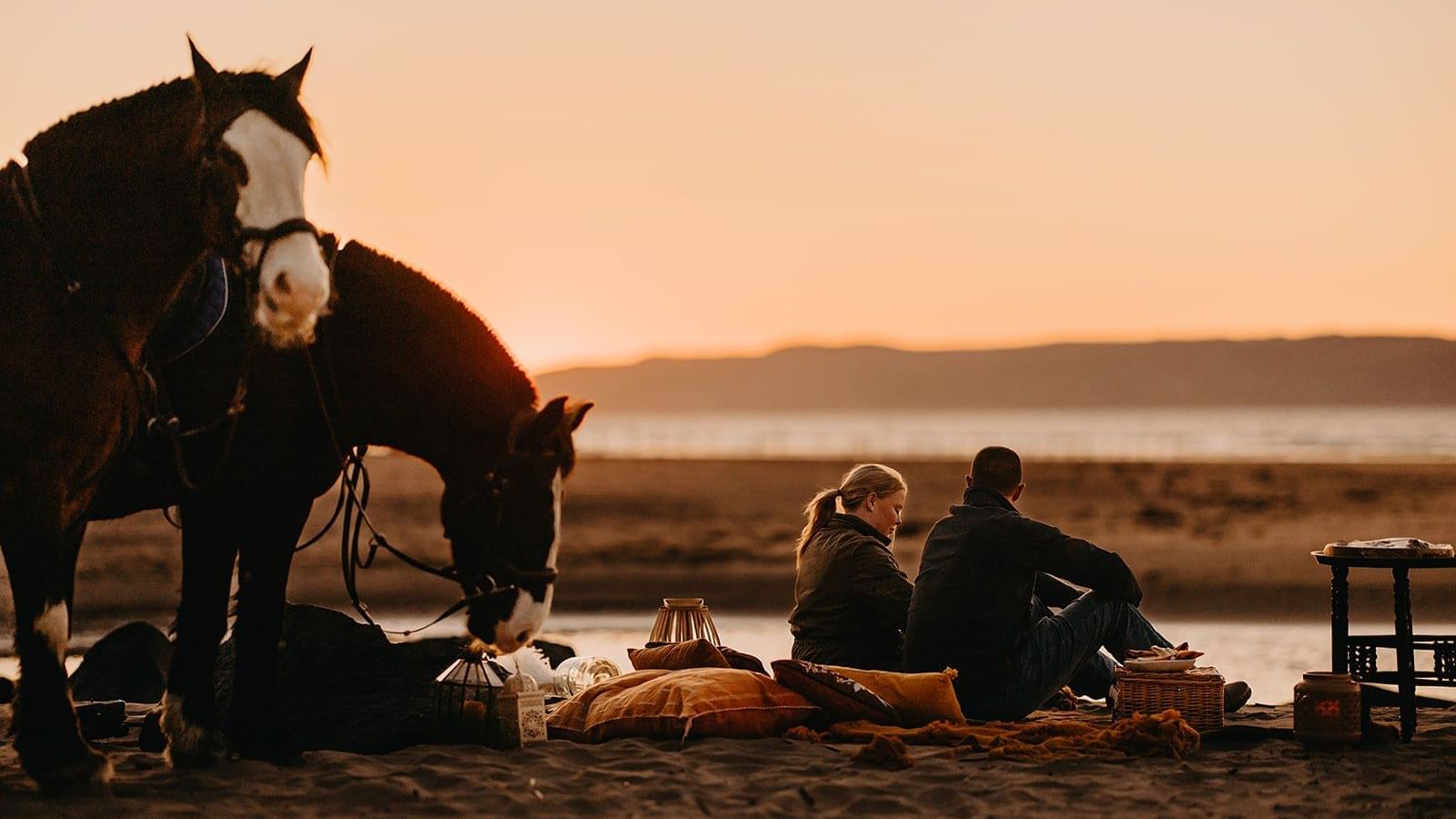 A couple sits on a beach during sunset with two horses nearby, enjoying a romantic picnic at Dunluce Lodge