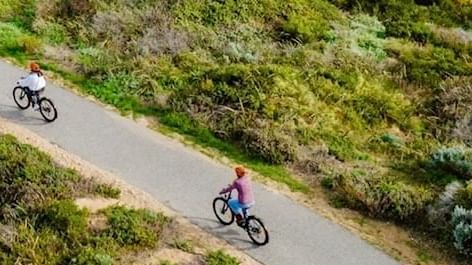 People ride bicycles along a path through a lush green landscape near The Sebel Mandurah