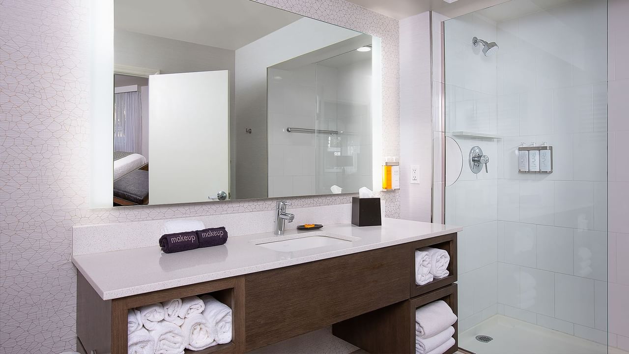 A bathroom with a large mirror, sink, and shower stall, featuring white towels and a makeup kit.