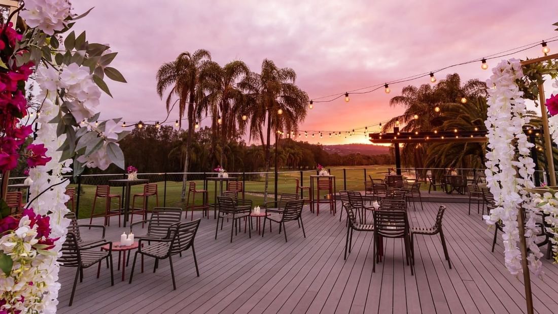 Decorated outdoor dining area with tables, chairs, string lights, and floral arrangements at sunset.