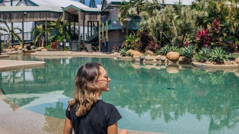 A person sits by a tropical pool, enjoying a drink, with lush greenery featuring Pool Pass at Mercure Hotel Townsville