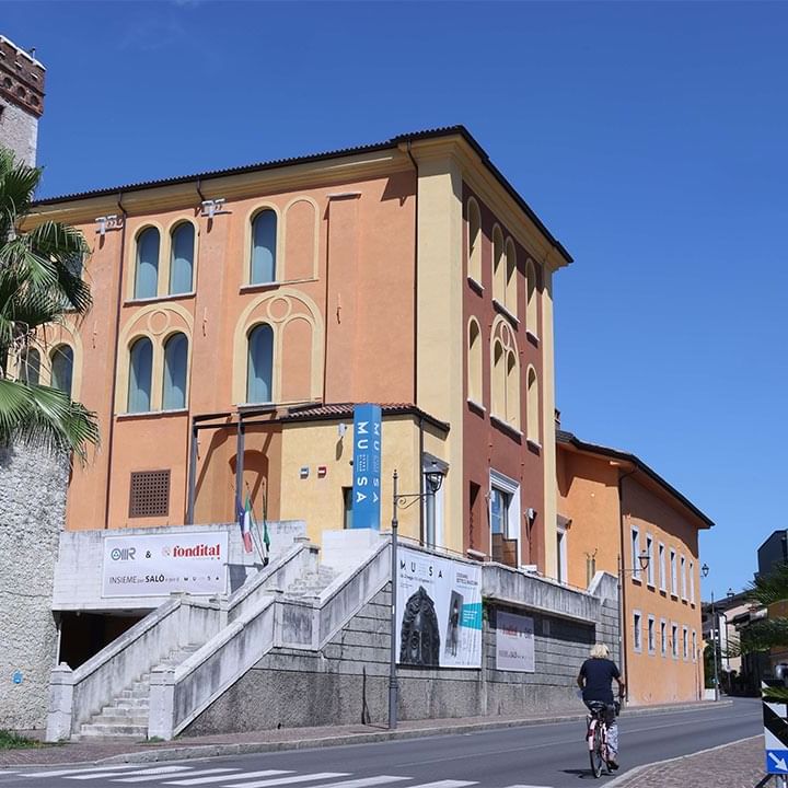 Ciclista su strada davanti a edificio storico con palme e cartelli pubblicitari sotto cielo blu.