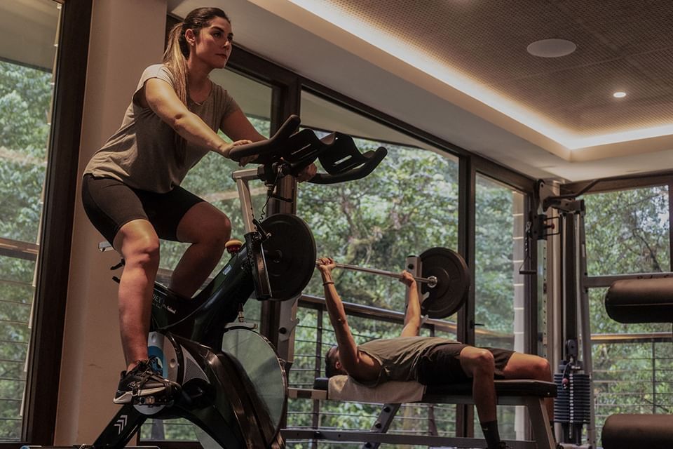 Two people exercising in a gym with large windows showing trees at El Silencio Lodge & Spa