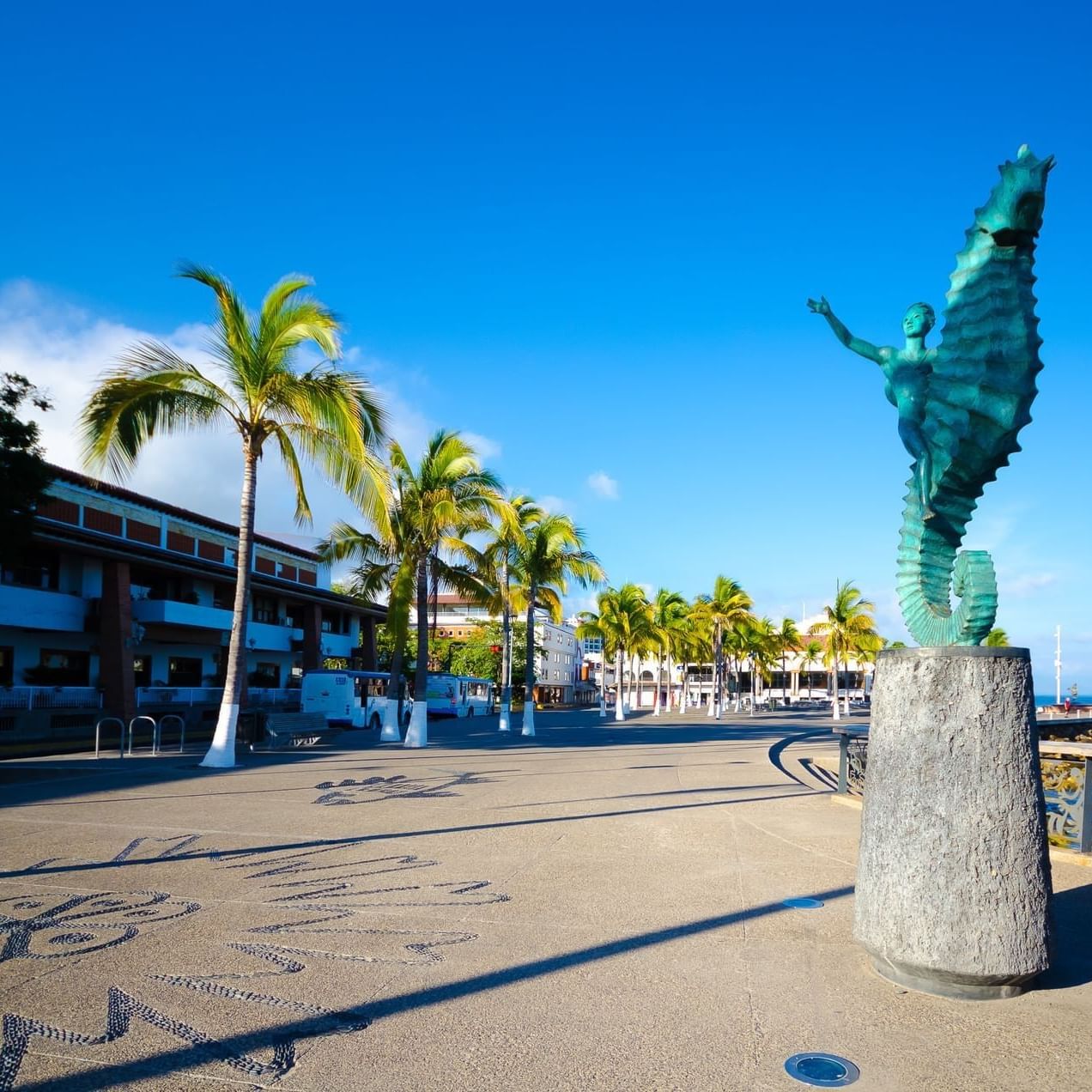 Estatua de una figura humana montando un caballito de mar en un paseo marítimo con palmeras y océano. El famoso caballito de Puerto Vallarta